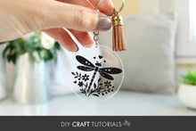 Load image into Gallery viewer, Hand holding a keychain with a dragonfly design and tassel, blurred background