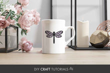 Load image into Gallery viewer, White mug with a dragonfly design on a wooden surface with flowers and decor in the background.
