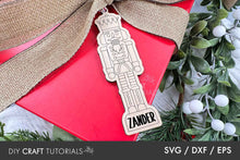 Load image into Gallery viewer, Decorative nutcracker tag with &#39;Zander&#39; on a red gift box surrounded by greenery.
