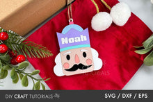 Load image into Gallery viewer, Decorative nutcracker ornament with &#39;Noah&#39; on a red fabric background with Christmas elements.
